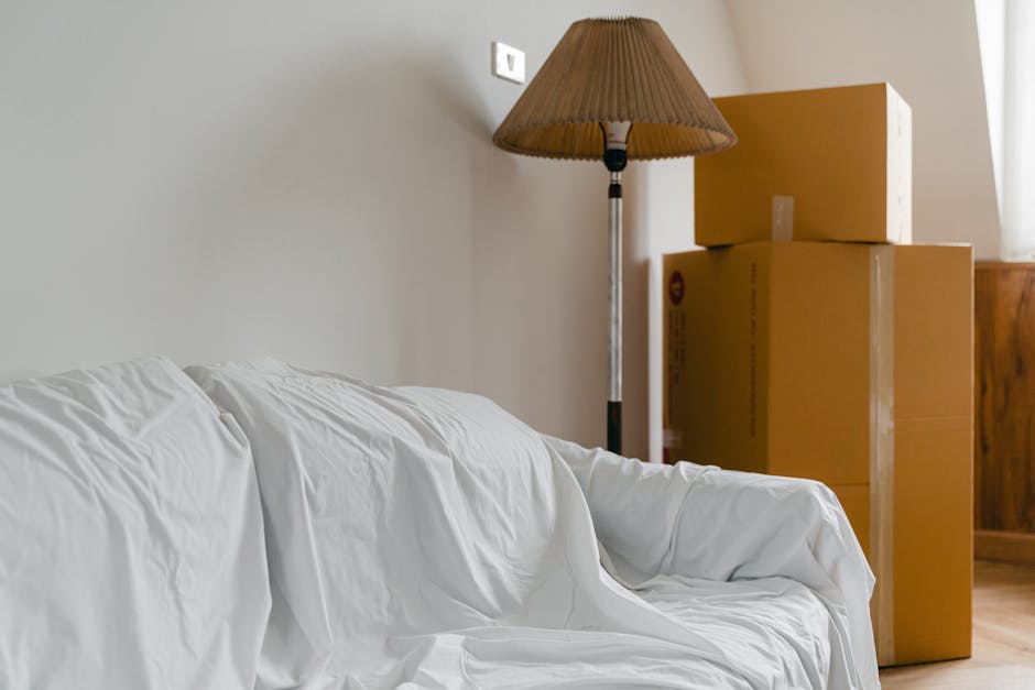 Inside a home indicating a home relocation scene, there are four cardboard moving boxes stacked in the corner near a wooden cabinet, with two larger boxes at the bottom and two smaller boxes on top. The boxes are sealed with packing tape and appear to contain household items. To the left, part of a white fabric-covered sofa is visible, with some crumples in the cushion cover. Behind the sofa, a tall floor lamp with a brown pleated lampshade stands, casting soft light onto the scene. The room has neutral-colored walls and wooden flooring, suggesting a typical moving environment. The setting implies a packing and moving process as part of a house removal service, supported by the presence of boxes ready for transport or loading. Man With a Van Enfield, a professional removals company, often handles such scenes during house relocations in the Enfield area, focusing on efficient furniture transport and packing logistics.