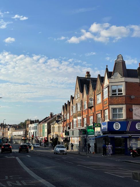The image depicts a busy urban street scene during daytime in Enfield, with a row of multi-storey brick and painted buildings lining the pavement. Several pedestrians are walking along the sidewalk, some near the entrance to a shop or service resembling a retail or convenience store. Parked and moving cars are visible on the road, with a few vehicles waiting at a traffic signal in the distance. The street features traffic lanes, pedestrian crossings, and various street signs, all illuminated by natural daylight under a bright, partially cloudy sky. The scene illustrates a typical area where house removals and furniture transport could take place, with the environment suitable for professional moving services that involve loading furniture, boxes, and packing materials into vans parked nearby, as coordinated by companies like Man With a Van Enfield.