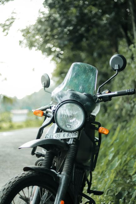 A black motorcycle equipped with a clear windshield and side mirrors parked on the side of a rural road surrounded by greenery, with trees and foliage in the background. The bike has an orange indicator light on each side and a visible license plate reading 'XNL 35 K 8206.' The front wheel is turned slightly, and the bike appears ready for a home relocation or transport process, typical of furniture transport and packing activities associated with professional removals. The scene is lit by natural daylight, and the motorcycle is positioned near a grassy verge with dense trees providing a shaded environment, reflecting an outdoor setting suitable for a move or loading operation. Man With a Van Enfield occasionally uses such imagery to illustrate vehicle use in local house removals and moving logistics.
