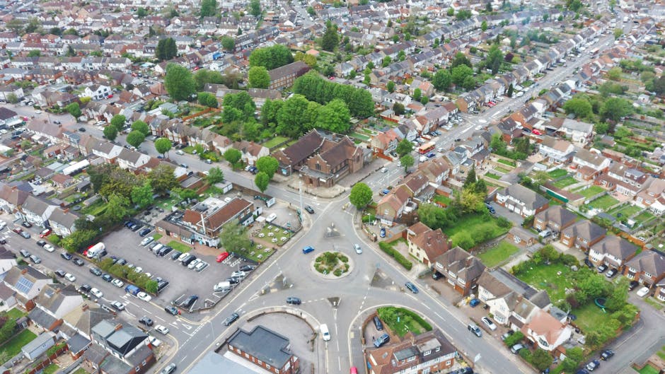 An aerial view of a residential neighbourhood in Enfield, featuring a roundabout with multiple entry and exit roads. Surrounding the roundabout are parked cars along the streets and in nearby parking areas, with terraced houses, semi-detached homes, and small gardens visible. Several houses have visible driveways, some with vehicles parked in them. Green trees line the streets and fill the front yards, providing a leafy backdrop. The image captures a typical home relocation environment, with cars possibly involved in loading or unloading, and the overall setting illustrating an area suitable for moving and furniture transport services. The image supports the context of house removals and relocation logistics offered by Man With a Van Enfield.