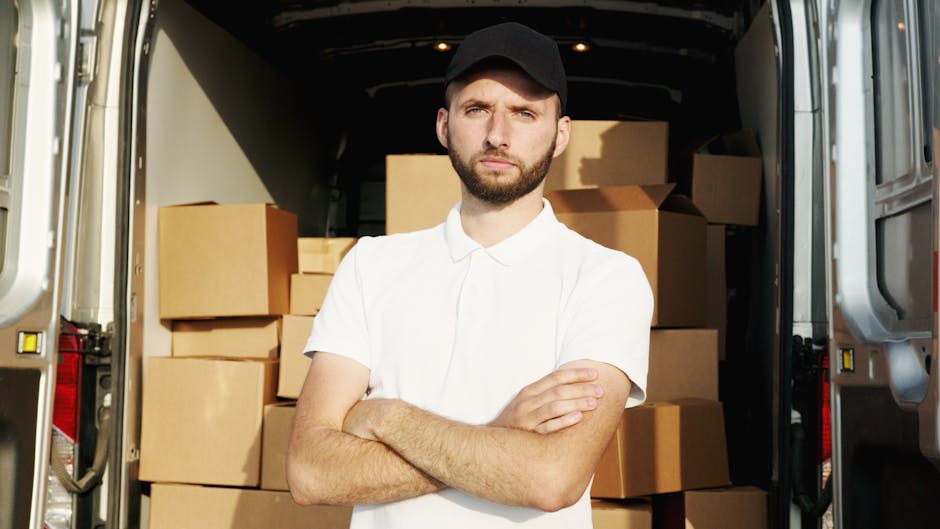 A man wearing a black cap and a light-colored polo shirt is inside a van with an open rear door, loading or arranging several cardboard boxes stacked on a wooden floor. The boxes vary in size, some ar