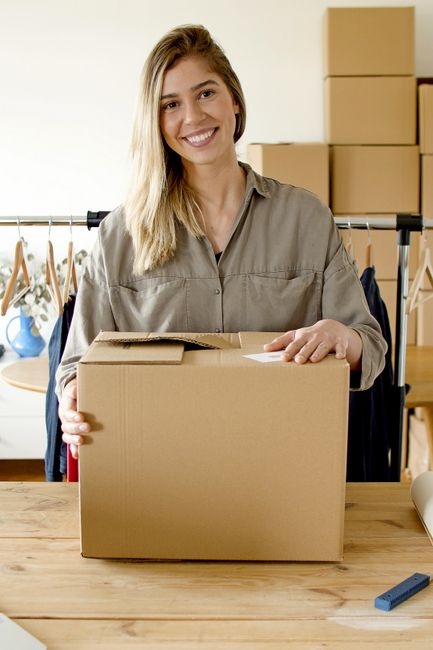 A young woman with long blonde hair and a wide smile is standing indoors behind a wooden table, holding a closed cardboard moving box with both hands. She is wearing a light grey collared shirt. In the background, there are several larger stacked cardboard boxes on a shelf or furniture unit, and a clothing rack with hangers and garments, indicating an environment related to packing or moving preparations. The lighting is bright and natural, suggesting a well-lit room, possibly during a house or office relocation process. The scene reflects organizing, packing, and preparing items for transportation, consistent with house removals or professional moving services offered by Man With a Van Enfield, especially relevant to the Oakwood and Cockfosters area.
