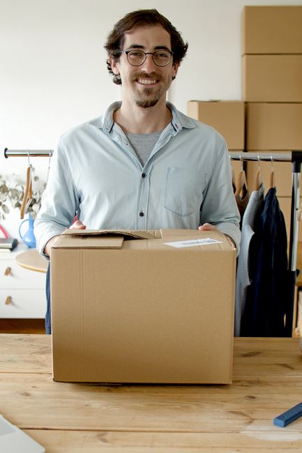 A man with glasses, dark wavy hair, and a light grey shirt is standing indoors, smiling at the camera while holding a medium-sized cardboard box with clear packing tape sealing the top. Behind him, an open wardrobe filled with hanging clothes and items, along with a clothing rack, indicates a domestic setting. To his right, on a wooden table, there are additional cardboard boxes and packing materials such as bubble wrap and plastic wrap, suggesting packing and moving preparations for an office or home relocation. Natural daylight illuminates the space, highlighting the organized environment typical of professional removal services. The scene reflects furniture transport and packing processes involved in house removals, and the professional approach of Man With a Van Enfield in managing small business or residential moves, aligning with the services referenced on the Oakwood and Cockfosters office removals page.