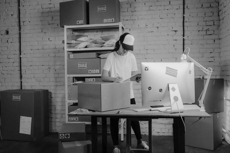 A young woman with long blonde hair and a wide smile is standing indoors behind a wooden table, holding a closed cardboard moving box with both hands. She is wearing a light grey collared shirt. In the background, there are several larger stacked cardboard boxes on a shelf or furniture unit, and a clothing rack with hangers and garments, indicating an environment related to packing or moving preparations. The lighting is bright and natural, suggesting a well-lit room, possibly during a house or office relocation process. The scene reflects organizing, packing, and preparing items for transportation, consistent with house removals or professional moving services offered by Man With a Van Enfield, especially relevant to the Oakwood and Cockfosters area.