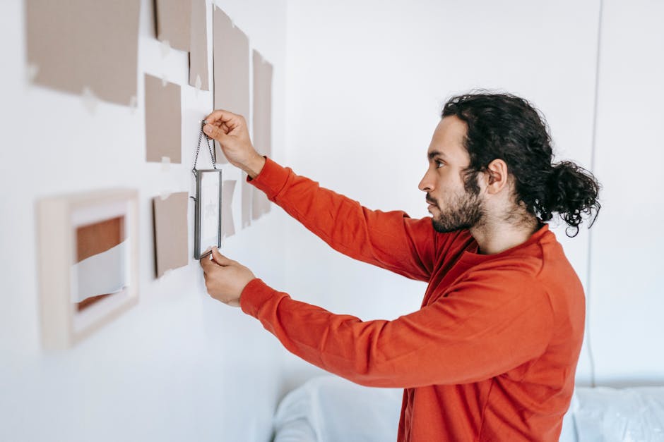 A man with dark, curly hair tied back in a ponytail, wearing a red long-sleeve shirt, is attaching a picture frame to a white wall that has several other frames and decorative items arranged in a gallery-style display. He is standing inside a well-lit room, possibly a living area or hallway. The man is carefully aligning the frame with his right hand, while his left hand holds the frame steady. The wall showcases a mix of small, square, and rectangular frames, some with images and others with empty backs or backing boards. The setting appears to be part of a home relocation process, with the man engaged in unpacking or decorating a new space, possibly assisted by Man With a Van Enfield, a company known for house removals and furniture transport services. The image captures the preparation and organization involved in packing and moving household items during a home relocation or home improvements.