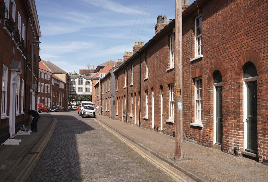 A quiet residential street with a cobblestone pavement in Enfield, featuring a row of red brick terraced houses with white-framed windows and doors. A man dressed in dark clothing is seen unloading or packing items on the sidewalk near the entrance of one of the houses, with white blankets or padding on the ground for protection. In the background, a white van is parked on the street, likely used for house removals or furniture transport by Man With a Van Enfield, whose services include home relocation and packing. A wooden utility pole stands along the pavement, with wires extending overhead. A few other parked cars are visible further down the street, and a modern building can be seen in the distance, connected by a small bridge or skywalk. The scene is well-lit with natural daylight, capturing the typical environment for local moving services and furniture loading processes.
