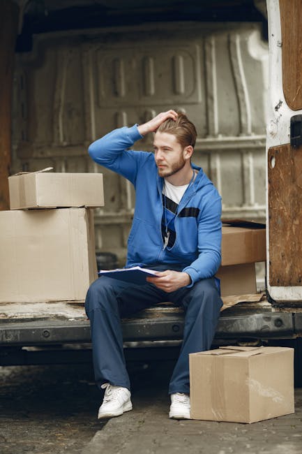 A young man with light brown hair and a beard, dressed in a blue jacket, navy trousers, and white sneakers, sits on the edge of an open delivery van during a home relocation. He holds a clipboard and appears to be taking notes or organizing the packing process. Surrounding him are several cardboard boxes, some stacked inside the van and others placed on the pavement nearby, indicating items awaiting loading or after unloading. The interior of the van has a metallic backing and is fitted with wooden panels, typical of commercial removal vehicles used by companies like Man With a Van Enfield. The scene is set outdoors on a paved surface, with natural lighting highlighting the workspace involved in furniture transport and packing during a house removal, reflecting professional moving services focused on efficient logistics and furniture loading procedures.