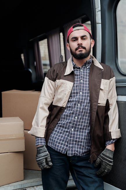 A man with a neatly groomed beard and dark hair, wearing a red backward cap, a checked shirt, and a beige and brown jacket, stands inside the back of a van used for house removals. The van's interior reveals several cardboard boxes, some open and others sealed, ready for transport. The boxes are stacked on the van's floor, with a few placed along the side walls, indicating a packing and loading process involved in home relocation. The lighting suggests natural daylight coming through the van's windows, which are partially visible. The man appears to be involved in a furniture transport or moving task, integral to professional removals services. Man With a Van Enfield is known for providing efficient loading and transportation services during house removals, ensuring safe and organized furniture and box handling.