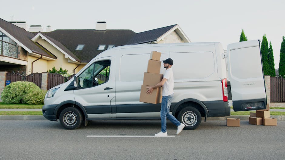 A young man with light brown hair and a beard, dressed in a blue jacket, navy trousers, and white sneakers, sits on the edge of an open delivery van during a home relocation. He holds a clipboard and appears to be taking notes or organizing the packing process. Surrounding him are several cardboard boxes, some stacked inside the van and others placed on the pavement nearby, indicating items awaiting loading or after unloading. The interior of the van has a metallic backing and is fitted with wooden panels, typical of commercial removal vehicles used by companies like Man With a Van Enfield. The scene is set outdoors on a paved surface, with natural lighting highlighting the workspace involved in furniture transport and packing during a house removal, reflecting professional moving services focused on efficient logistics and furniture loading procedures.